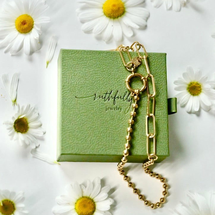 A gold chain necklace with paperclip and ball links rests on a textured light green jewelry box. The box has the brand name 'ruthfully jewelry' printed on it.